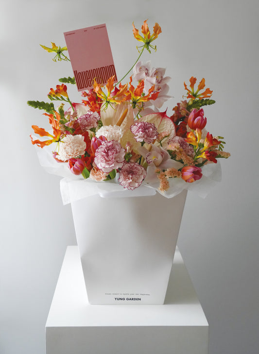 白色紙袋內裝着鮮艷明亮的母親節花束，由不同顏色的康乃馨為主花、配搭線條活潑的西施百合和鬱金香。花束插着粉紅色的母親節卡。