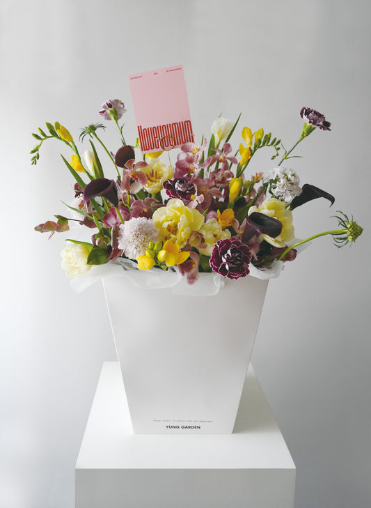 白色紙袋內裝着優雅美麗的母親節花束，由米黃色鬱金香為主花、配搭優雅的紫色蘭花、康乃馨和高貴的馬蹄蘭。花束插着粉紅色的母親節卡。