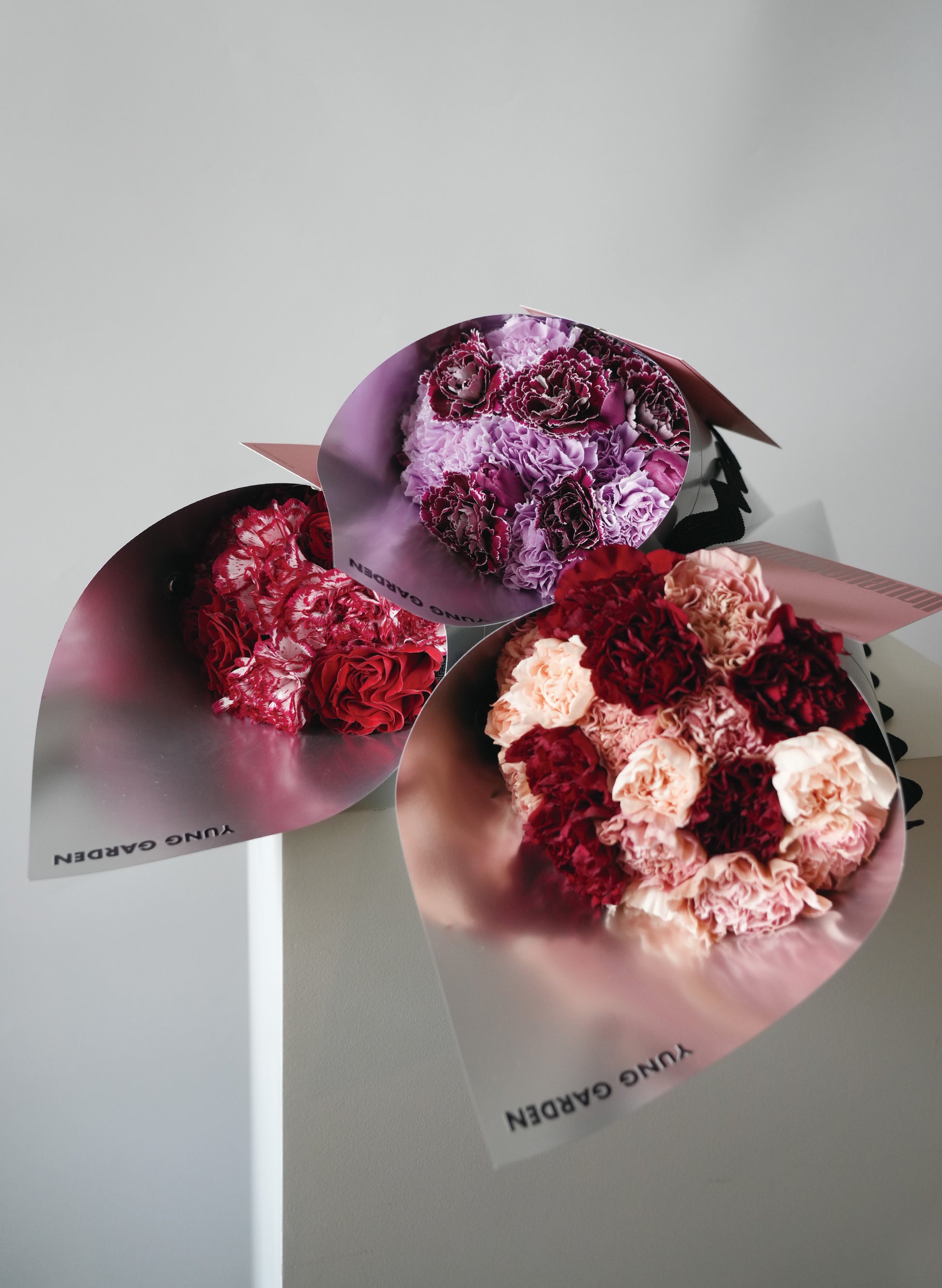 三個母親節花束疊在一起，每個都不同顏色，分別是紫色、紅色、和混合粉色。全都包裹在簡约的銀色印有"YUNG GARDEN"牌子名稱的包裝纸中。
