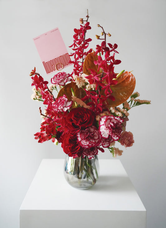 透明玻璃樽中有紅玫瑰、康乃馨、蘭花和其他襯花，左上角插着一張粉紅色的母親節咭。