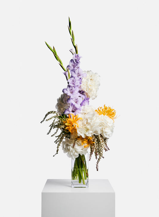 A beautiful seasonal flowers bouquet featuring hydrangea, gladiolus, lycoris sprengeriand in a clear glass vase. It is summer colour like yellow, white and pastel purple. The florist arrange them in a unique and cool shape. 